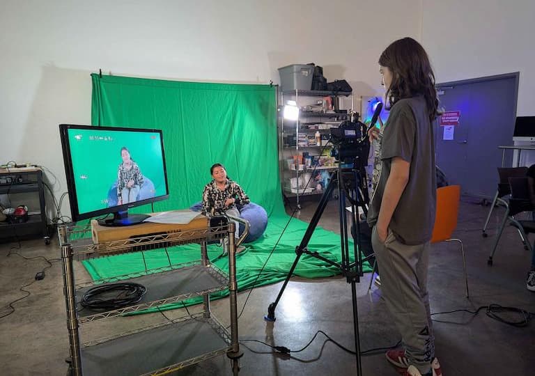 Students film in front of a green screen, seated on a bean bag.